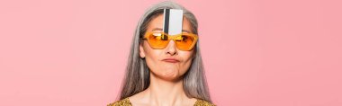 mature asian woman in yellow sunglasses with credit card near forehead isolated on oink, banner clipart