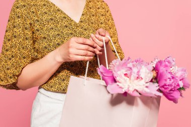 partial view of woman in stylish blouse holding shopping bag with peonies isolated on pink clipart