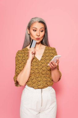 Kredi kartı ve cep telefonu tutan düşünceli Asyalı kadın pembeye izole edilmiş.