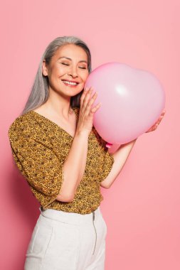 Mutlu Asyalı kadın, gözleri kapalı pembe arka planda kalp şeklinde balon tutuyor.