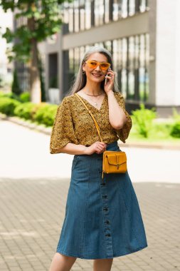 Gülümseyen Asyalı kadın modaya uygun bluz ve kot etek giyiyor. Şehir caddesinde akıllı telefondan konuşuyor.