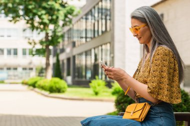 şık güneş gözlüklü olgun Asyalı kadın bankta oturuyor ve cep telefonuyla mesajlaşıyor.