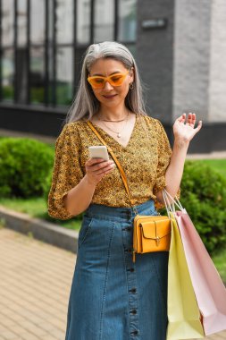 Çapraz vücutlu ve alışveriş çantalı şık Asyalı kadın şehir caddesinde akıllı telefon kullanıyor.