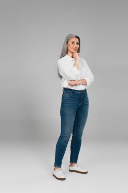 Beyaz tişörtlü ve kot pantolonlu Asyalı bir kadın. Gri arka planda çenesinin yanında duruyor.