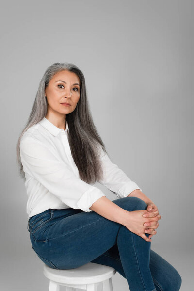 grey haired asian woman in white shirt and jeans sitting on stool isolated on grey