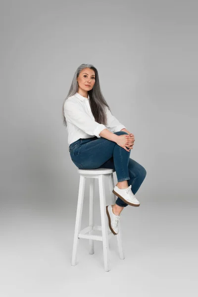 grey haired asian woman in white shirt and blue jeans looking at camera on high stool on grey background