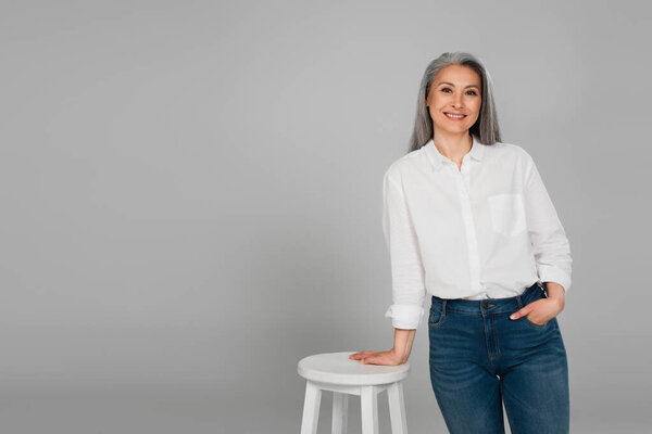 smiling asian woman in white shirt standing near high stool with hand in pocket of jeans isolated on grey