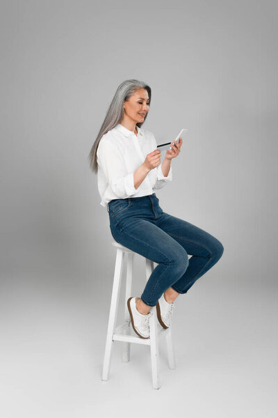 grey haired asian woman in jeans and shirt sitting on high stool with cellphone and credit card on grey background