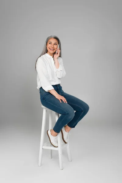 Alegre Asiático Mujer Jeans Sentado Alto Taburete Hablando Smartphone Gris — Foto de Stock