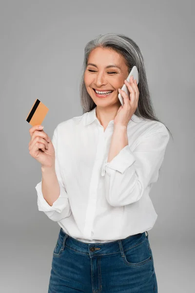 cheerful asian woman with credit card talking on cellphone isolated on grey
