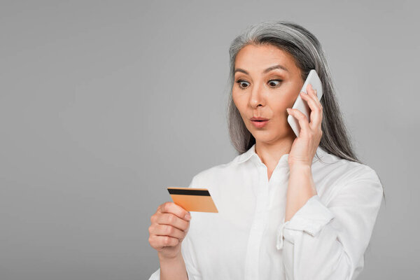 surprised asian woman calling on mobile phone while looking at credit card isolated on grey