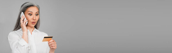 amazed asian woman looking at credit card while calling on cellphone isolated on grey, banner