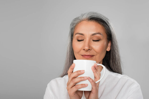 pleased asian woman enjoying flavor of tea isolated on grey