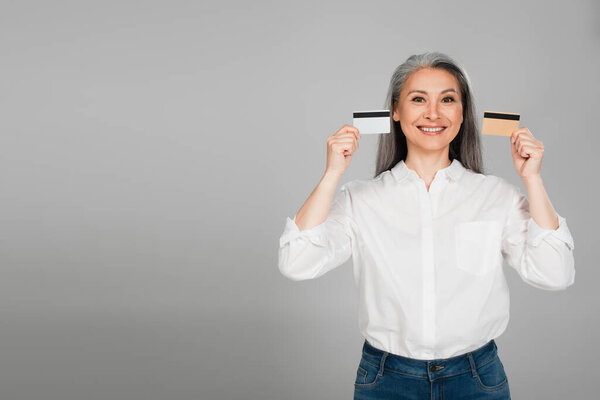 радостная азиатская женщина в белой рубашке с кредитными картами, изолированными на сером