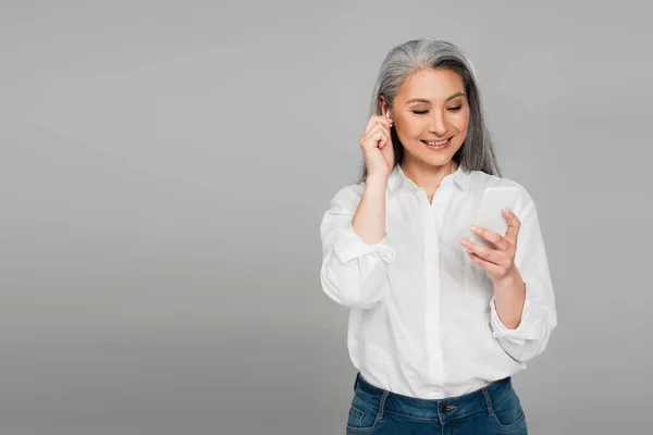 Alegre Asiático Mujer Ajuste Auricular Mientras Usando Smartphone Aislado Gris — Foto de Stock