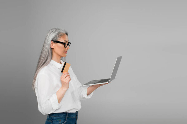 middle aged asian woman in white shirt and eyeglasses holding laptop and credit card isolated on grey