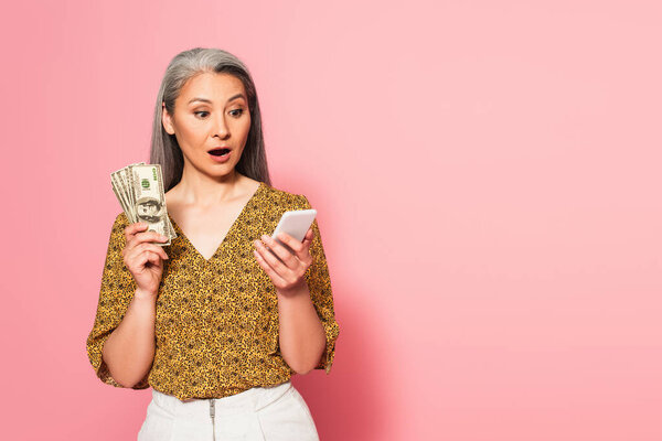 удивлена азиатская женщина с долларовыми банкнотами, глядя на смартфон на розовом фоне