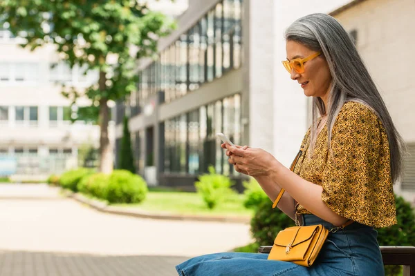 成熟的亚洲女人 戴着时髦的太阳镜 坐在长椅上 用手机发短信 — 图库照片