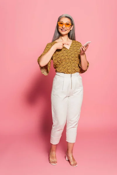 Volwassen Aziatische Vrouw Stijlvolle Kleding Zonnebril Wijzend Smartphone Roze Achtergrond — Stockfoto