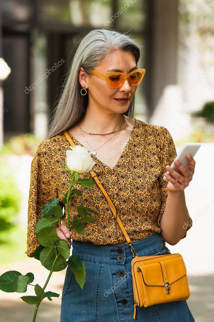 middle aged asian woman in trendy clothes holding white rose while messaging on smartphone outdoors