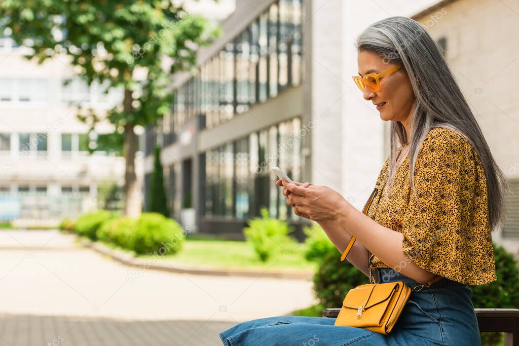 mature asian woman in stylish sunglasses sitting on bench and messaging on mobile phone