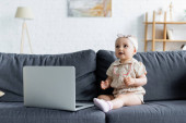 Dítě batole se dívá pryč poblíž notebooku na gauči 