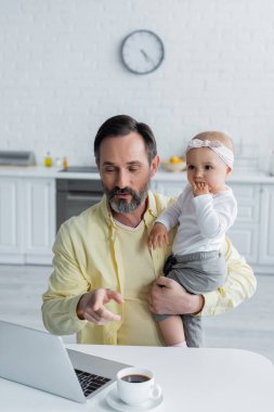 Yetişkin bir adam, bebek kızını mutfakta tutarken dizüstü bilgisayarı işaret ediyor. 