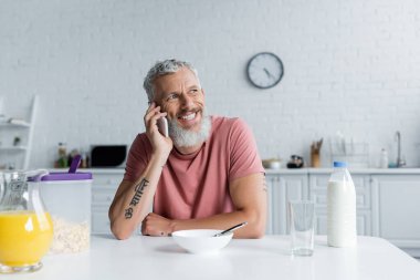 Mutfakta kahvaltıda akıllı telefondan konuşan gülümseyen olgun bir adam. 