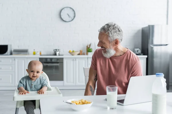 Lächelnder Vater Schaut Baby Auf Hochstuhl Neben Frühstück Und Laptop — Stockfoto