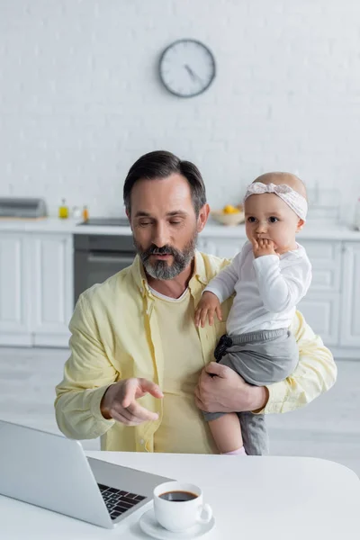 Yetişkin Bir Adam Bebek Kızını Mutfakta Tutarken Dizüstü Bilgisayarı Işaret — Stok fotoğraf
