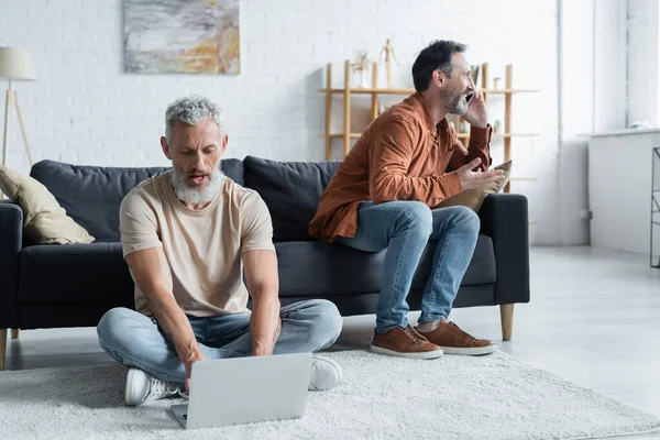 Homosexual Hombre Usando Portátil Cerca Pareja Hablando Teléfono Inteligente Casa —  Fotos de Stock