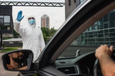 Tehlikeli madde giysisi giymiş bir tıp çalışanı arabada bulanık bir sürücünün yanında el sallıyor. 