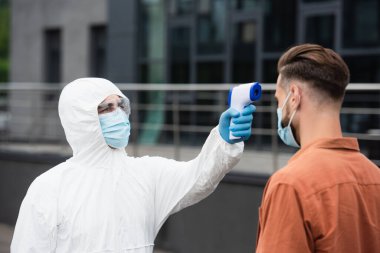 Tehlikeli madde giysisi giymiş bir tıp çalışanı açık havada maske takan bir adamın sıcaklığını ölçüyor. 