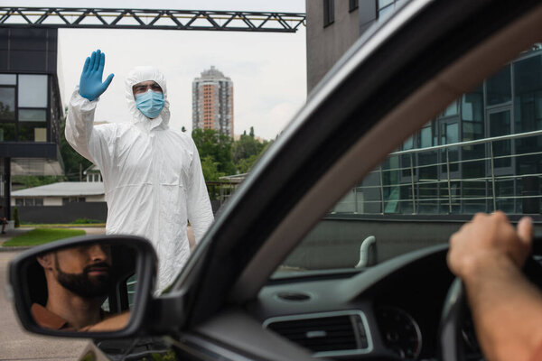 Medical worker in hazmat suit waving hand near blurred driver in car 