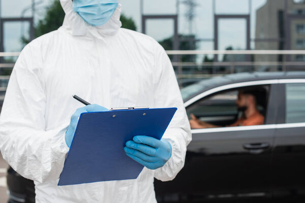 Medical worker in hazmat suit writing on clipboard near blurred man in car 