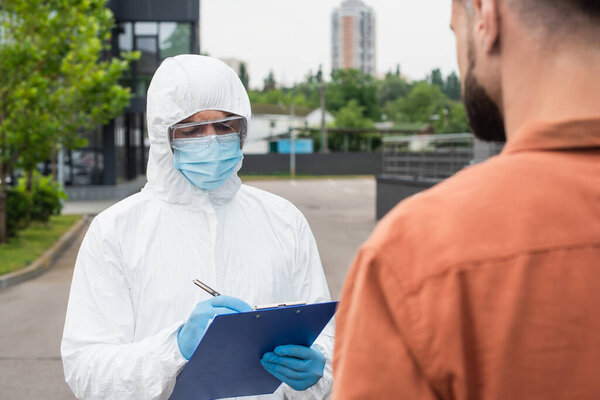 Medical worker in hazmat suit writing on clipboard near blurred man outdoors 