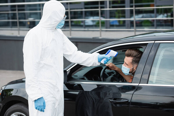 Medical worker in protective suit holding pyrometer near man in mask in car 