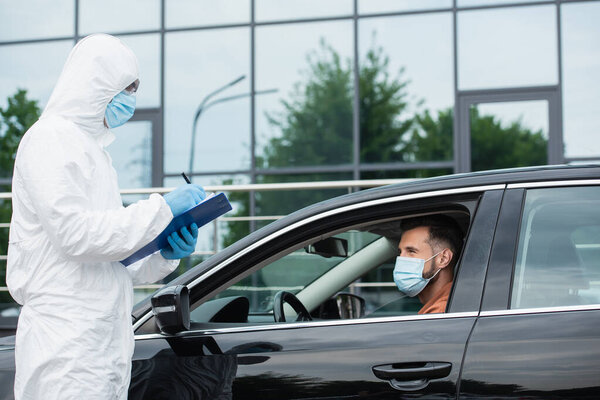 Medical worker in hazmat suit writing on clipboard near driver in medical mask 