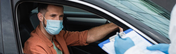 Driver in mask looking at blurred medical worker writing on clipboard, banner 