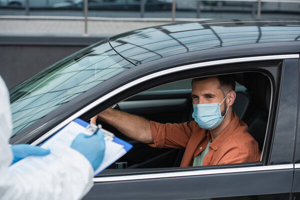 Driver in mask looking at medical worker in latex gloves writing on clipboard 