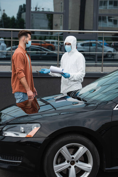 Driver in mask standing near medical worker with clipboard pointing with hand outdoors 