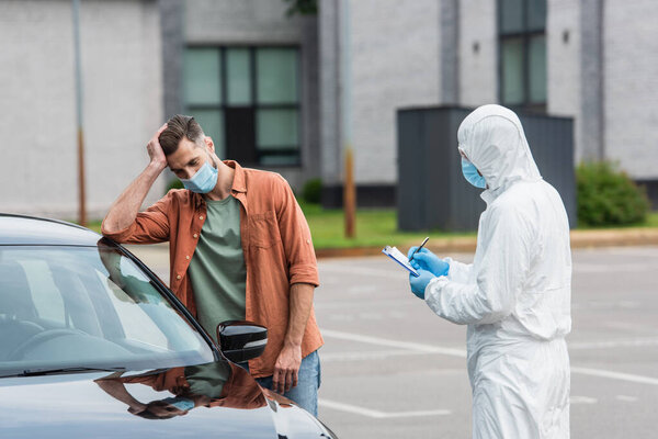 Sad driver standing near medical worker in hazmat suit writing on clipboard 