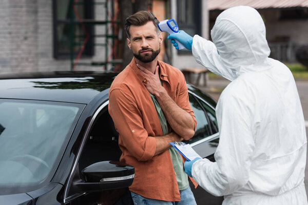 Ill driver standing near medical worker with clipboard and pyrometer outdoors 