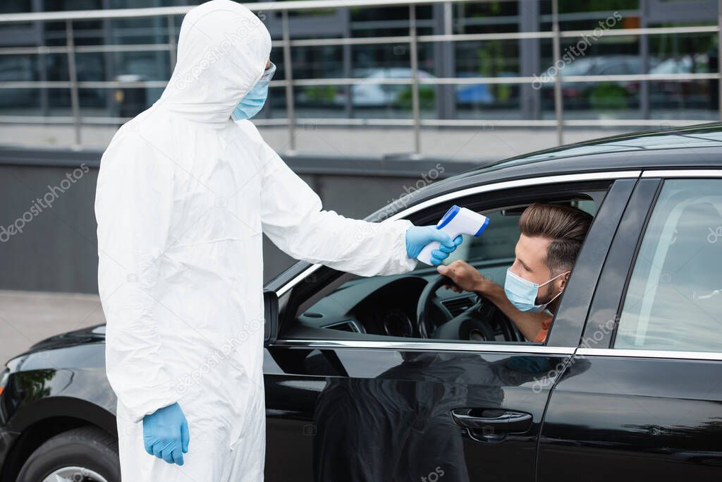 Medical worker in protective suit holding pyrometer near man in mask in car 