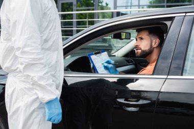 Sağlık çalışanının yanında tehlikeli madde kıyafeti giymiş bir sürücü panosunda yazıyor. 