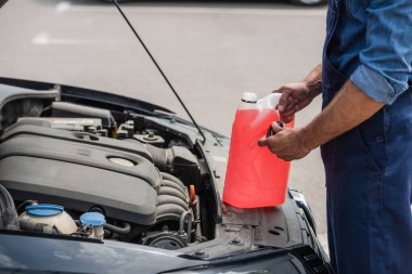 Cropped view of mechanic holding canister of windshield washer fluid and cap near car clipart