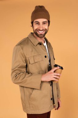 cheerful man in brown beanie and jacket standing with coffee to go isolated on beige