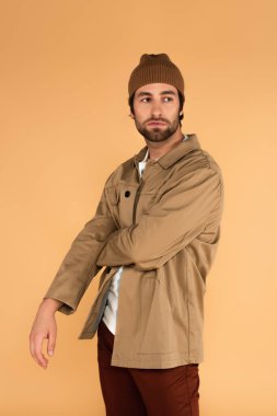 young man in jacket and brown beanie looking away while standing isolated on beige