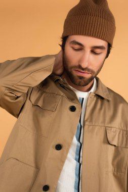 trendy man in brown beanie and jacket posing with hand on neck isolated on beige