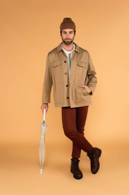 man with hand in pocket of trendy jacket leaning on folded umbrella on beige